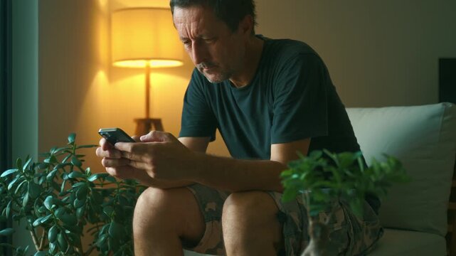 Middle-aged man sits on a sofa in a cozy living room, focused on his smartphone in the evening light. A calm, solitary moment of technology, routine and quiet reflection at home. 4K.
