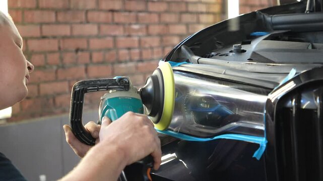 Man polishes the glass of a car headlight with a polishing machine from small scratches, chips. Process polishing of the front car headlights. Hands of male professional worker with orbital polisher. 