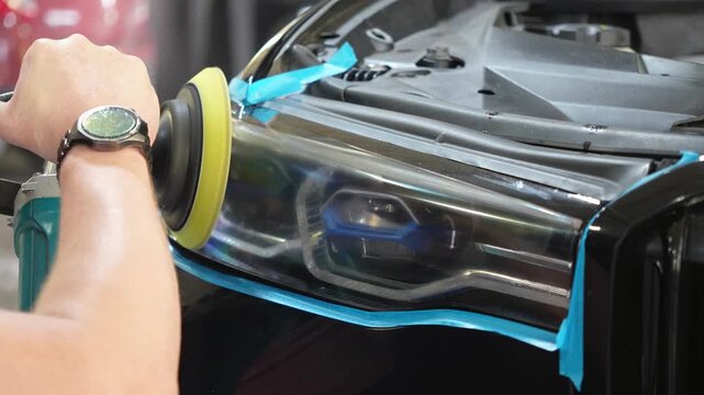 Man polishes the glass of a car headlight with a polishing machine from small scratches, chips. Process polishing of the front car headlights. Hands of male professional worker with orbital polisher. 