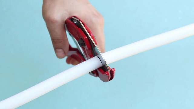 The process of cutting a white plastic pipe with a red pipe cutter. The master uses a manual ratchet pipe cutter to cut a PVC pipe on a blue background. Close-up of a tool in operation