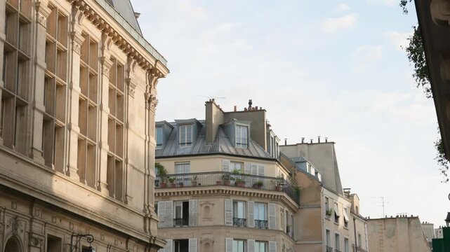 Architectural view of historical Parisian street buildings featuring detailed stone facades, window shutters, and a zinc mansard roof under a bright sky. Camera tilting up