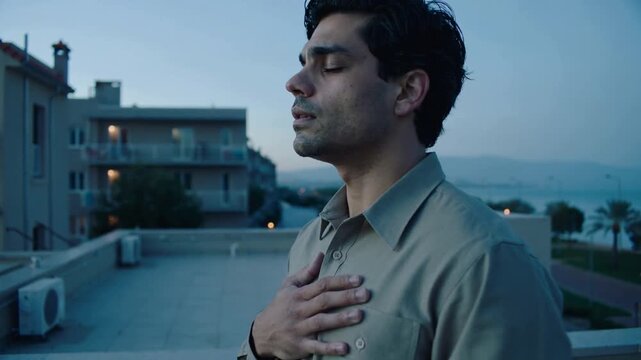 Inhaling man in collared shirt with right hand on chest, tilting back, calming on rooftop parapet