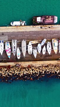 Sailboat fleet docked at stone pier, Nesebar, Bulgaria. Top down aerial view of white sailboat hull, cargo barge and turquoise harbor water. Vertical video.