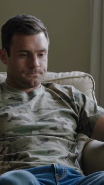 Vertical video: Gazing down, man in camo tee leaning forward on couch at home, showing concern