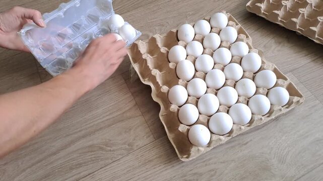Top view of hands transferring white chicken eggs from a large cardboard crate into a smaller plastic carton on a wooden floor background.