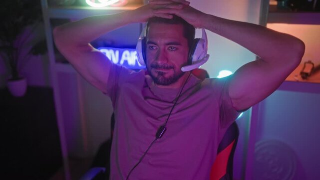 Man with headset grips head with both hands in a streaming studio with neon on air sign and gaming chair visible; frustration defeat.