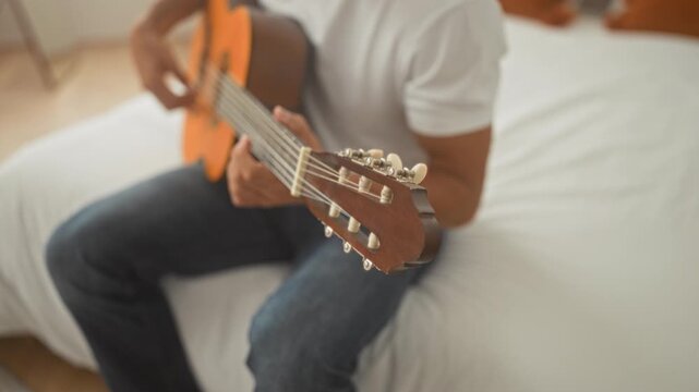 Man strumming acoustic guitar on bed in studio, hands fingering chords and focused expression while seated on white bedding and orange cushions; concentration.