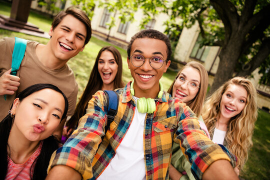 Happy diverse group of college students and friends taking a selfie together on a sunny campus green, laughing and enjoying college life
