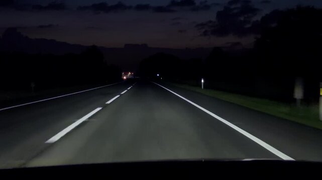 Night highway drive with empty lanes, distant headlights, dark trees, and moody sky over rural road
