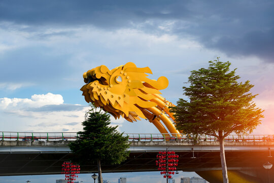 The striking golden dragon head of Da Nang's Dragon Bridge stands over the Han River, flanked by green trees and decorative red lanterns under a cloudy sky