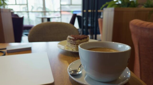 Coffee cup and layered dessert on wooden table in bright cafe lounge with notebook and soft blurred background