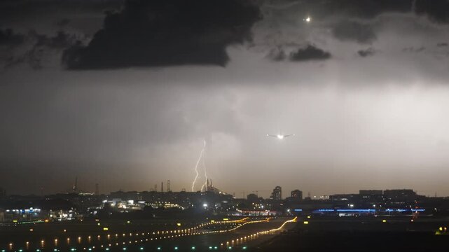 Airplane on final approach at night with bright landing lights as a lightning bolt strikes over the distant city skyline, runway lights glow along the airport in slow motion