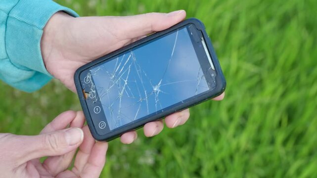 Person holding a smartphone with a broken screen. Close up of a person's hands holding a modern mobile phone with a cracked and shattered screen after being dropped, showing the damage