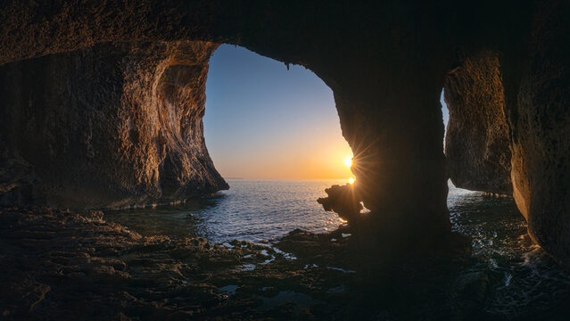 Dark rocky cave frames a golden sunrise over the calm sea along the Golfo di Orosei coast.