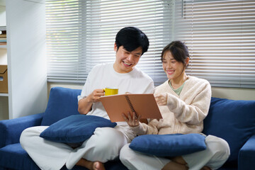 Fototapeta premium Young happy couple sits together on a cozy blue couch in their living room, looking at a memory book together and smiling while reminiscing about cherished times and drinking coffee.