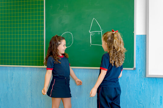 Young girls in school uniform looking at each other, drawing shapes on a blackboard during a class creating friendship and learning