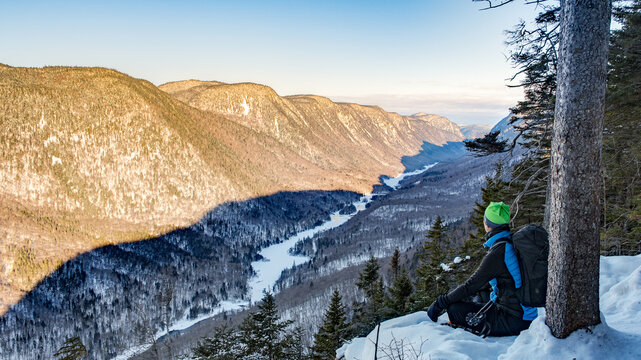 Lone female hiker sitting at the edge of the snowy valley at dawn, Jacques-Cartier national park in winter, Quebec, Canada