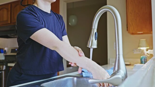 A person stands at a kitchen sink, washing their hands under running water. They use a cloth nearby and are focused on cleanliness while in their home kitchen.