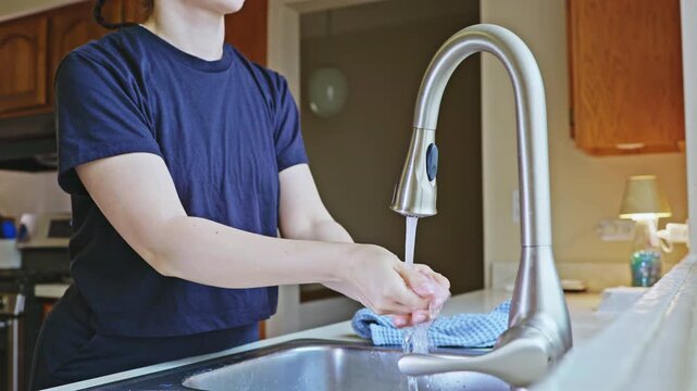 A person stands at a kitchen sink and washes hands with water flowing from a faucet. The kitchen appears simple and functional for daily tasks. A towel lies nearby.