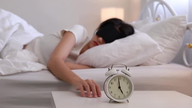 A sleepy young woman with an eye mask is turning off the alarm clock on her bedside table. She presses the alarm button, waking up to the sound of the alarm while she was sleeping soundly.