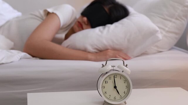A sleepy young woman with an eye mask is turning off the alarm clock on her bedside table. She presses the alarm button, waking up to the sound of the alarm while she was sleeping soundly.