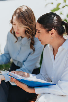 Two businesswomen sit together reviewing papers and a blue folder while discussing work. The colleagues collaborate in an outdoor meeting, examining documents and taking notes.