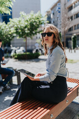Fototapeta premium A businesswoman sits on a city bench using a tablet while wearing sunglasses. The professional works remotely in an urban outdoor setting, focused and composed.