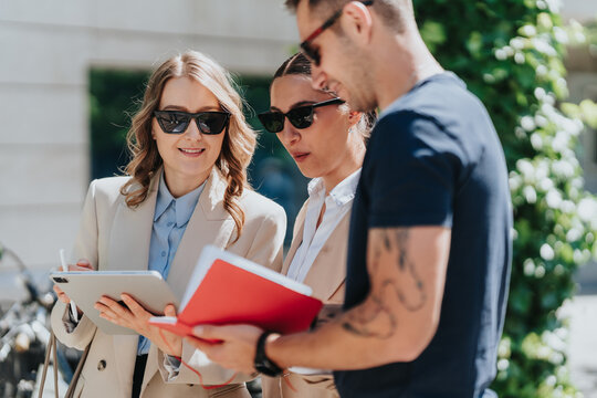 Three young professionals review a red folder and digital tablet outdoors during a business meeting. Colleagues stand together consulting documents and a tablet in a casual work setting.