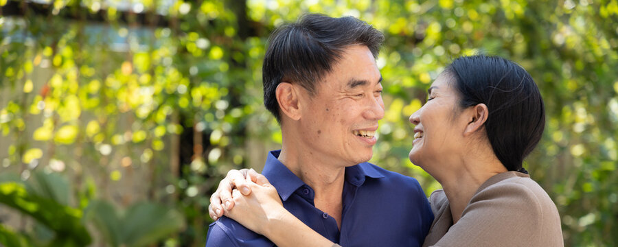 Happy smiling lovely Asian senior couple, elderly wife hugging her husband in urban park with good quality of life, concept photography of old aged togetherness and senior lovers intimacy, header crop