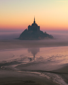 Aerial view of the historic Mont Saint-Michel abbey rising above the tidal flats and thick fog during a soft sunset in Mont Saint-Michel, Normandy, France.
