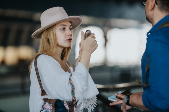 A female tourist adjusts her makeup using a compact mirror while waiting in a transit area. A nearby traveler holds travel documents as they prepare to continue their journey.