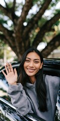 Fototapeta premium Smiling young woman waving from car window for lifestyle portrait and social media banner and greeting imagery for everyday urban content