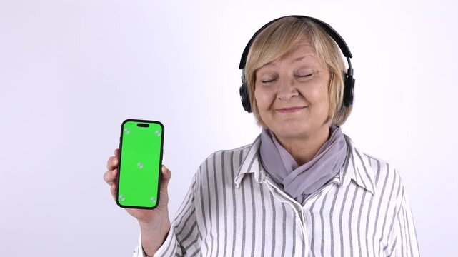 Senior woman wearing wireless headphones and holding a smartphone with green screen for mobile app, music streaming or audio service mockup.
