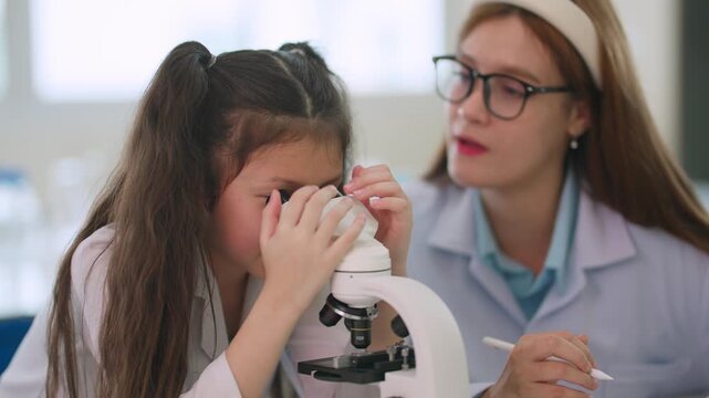 Young girl peers through microscope with curiosity. Teacher watches closely, guiding her with gentle care. Scientific discovery begins in bright lab setting. Perfect for educational, research