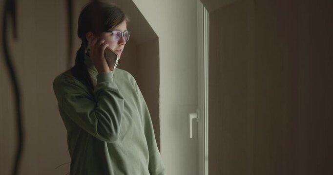 Thoughtful Girl Talks on Phone Looking out of Window. 4K 10-bit