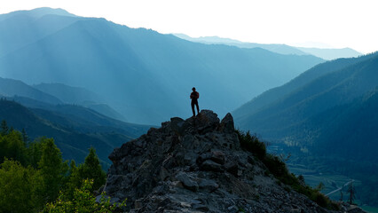 An athletic male tourist standing on top of a rocky cliff at Chike-Taman Pass, looking at his smartphone against a majestic blue mountain valley background. © OlTarakanov