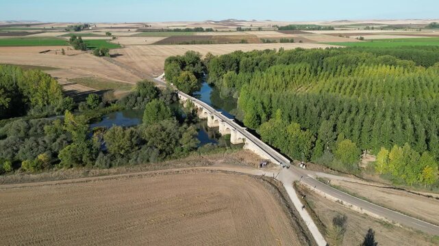 An aerial view of a rural bridge crossing a calm river beside dense tree rows and wide agricultural fields along the Camino de Santiago in Spain