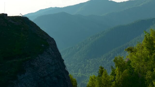 A cinematic telephoto drone rising shot from behind a dark rocky cliff, revealing a majestic panorama of misty mountain ranges and distant green valleys at sunrise.