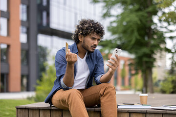Obraz premium Stressed young indian man sitting outdoors, holding a credit card and looking at his smartphone with a confused and worried expression while dealing with online banking issues