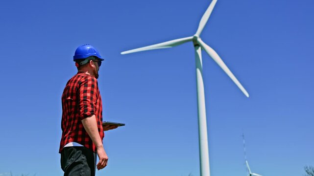 A windmill engineer stands in front of a wind turbine looking at a tablet. The worker is in a field with blue sky above. It is a bright day and the turbine blades are spinning.