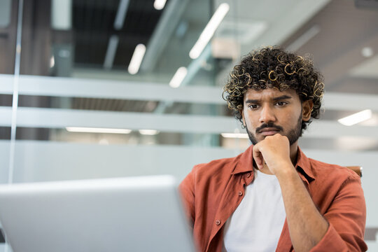 Concentrating young man with curly hair intently looking at a laptop screen in a modern office, deep in thought while planning a business project or analyzing data