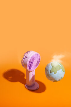 Minimalist global warming concept. Steaming Planet Earth sphere overheating on a plain orange background, facing a running electric desk fan in an attempt to cool down with copyspace