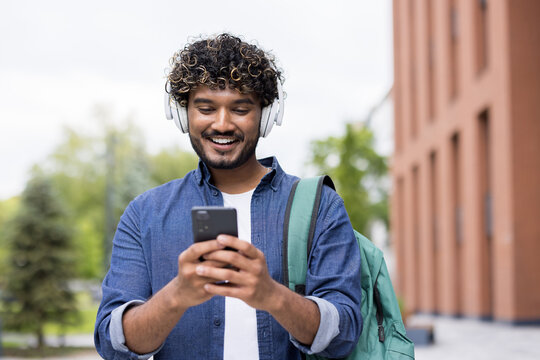 Happy indian student wearing headphones, actively using a smartphone, and carrying a backpack while walking on a university campus, representing modern higher education and connectivity