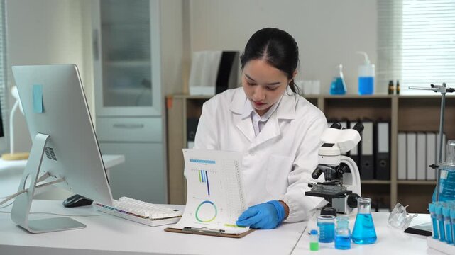 Asian female scientist analyzing data on computer and clipboard in modern laboratory, doing research and development of new drugs and medical treatments for biotechnology