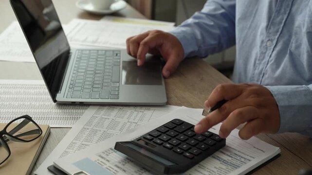 man Hands review financial reports with a calculator and pen beside a laptop, checking figures on printed documents for accounting analysis, budgeting records, and business finance workflow.