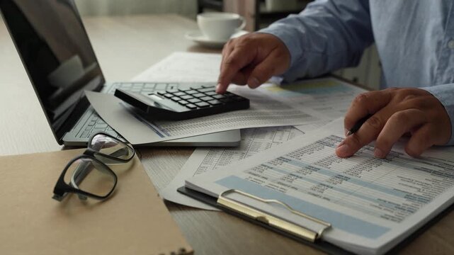 man presses a calculator over accounting reports beside a laptop, reviewing financial figures with a pen on a desk for report preparation, bookkeeping tasks, and business finance content.
