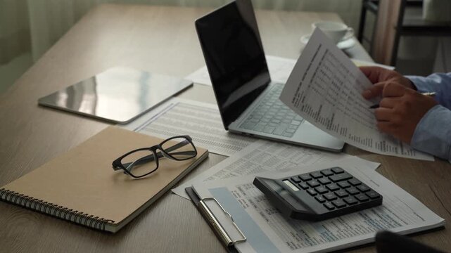 man presses a calculator over accounting reports beside a laptop, reviewing financial figures with a pen on a desk for report preparation, bookkeeping tasks, and business finance content.