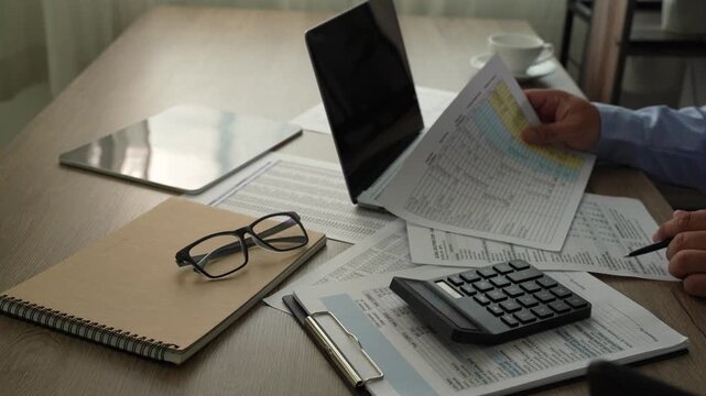 man Hands review financial reports with a calculator and pen beside a laptop, checking figures on printed documents for accounting analysis, budgeting records, and business finance workflow.