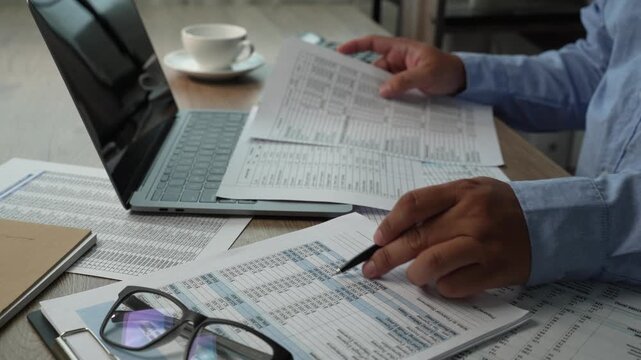 man presses a calculator over accounting reports beside a laptop, reviewing financial figures with a pen on a desk for report preparation, bookkeeping tasks, and business finance content.