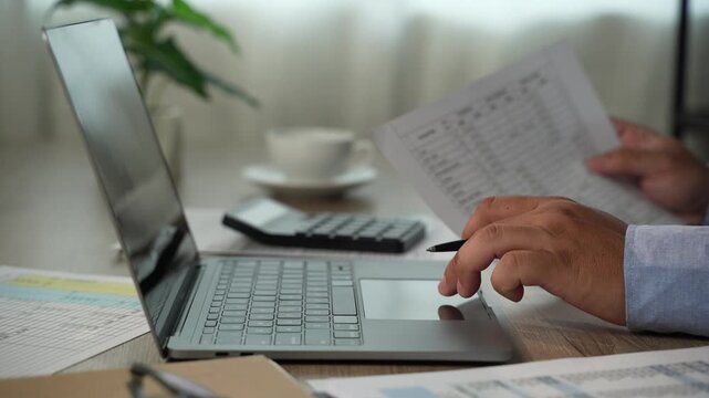man presses a calculator over accounting reports beside a laptop, reviewing financial figures with a pen on a desk for report preparation, bookkeeping tasks, and business finance content.